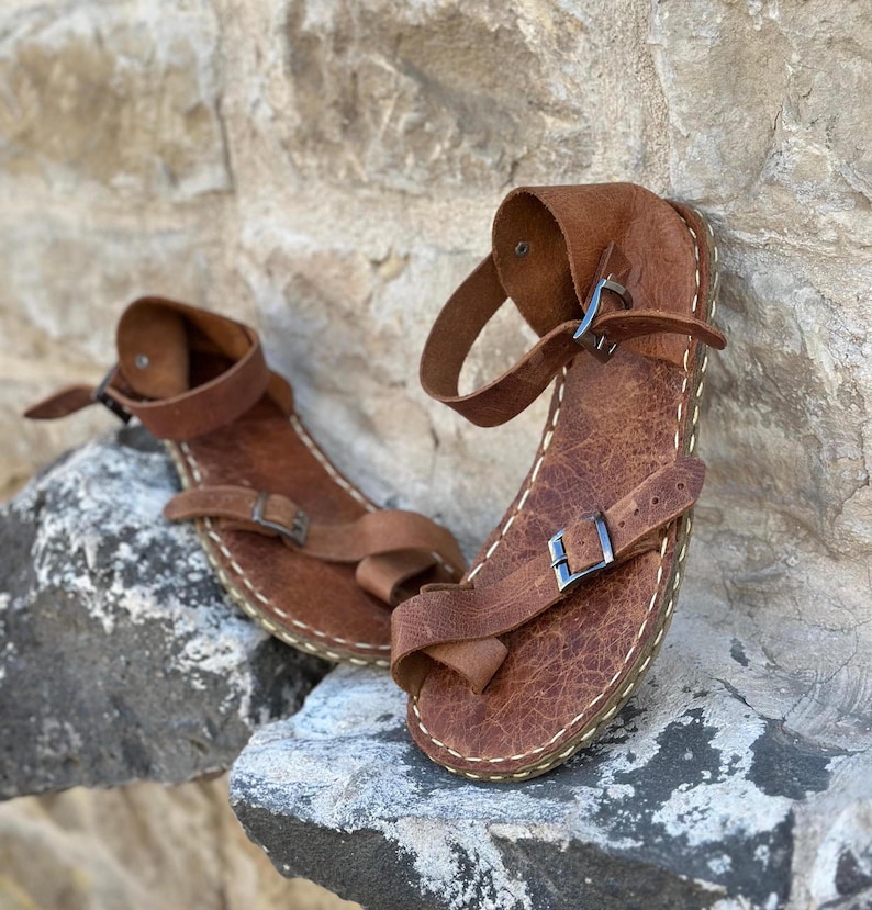 May include: A pair of brown leather sandals with buckles and straps. The sandals have a stitched edge and are sitting on a rocky surface.