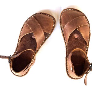 Op de afbeelding: Een paar bruinleren sandalen met een kruisvormig ontwerp over de voet en een enkelbandje. De sandalen hebben witte stiksels rond de randen en een gespsluiting. Getoond op een witte achtergrond.