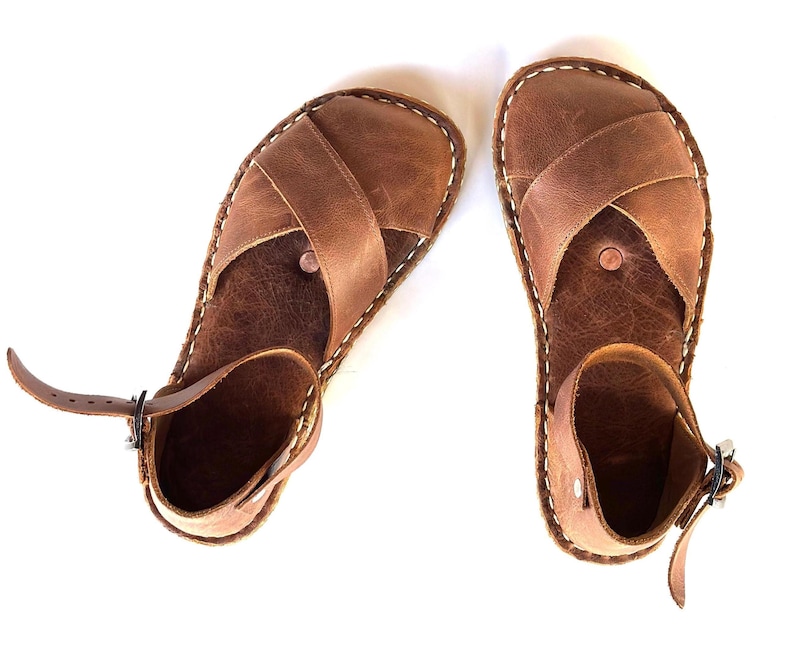 Op de afbeelding: Een paar bruinleren sandalen met een kruisvormig ontwerp over de voet en een enkelbandje. De sandalen hebben witte stiksels rond de randen en een gespsluiting. Getoond op een witte achtergrond.