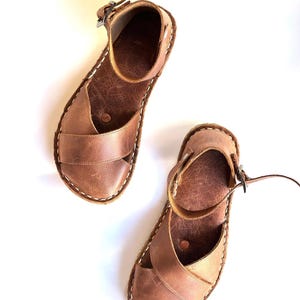 Op de afbeelding: Een paar bruine leren sandalen met een bandje en gespsluiting. De sandalen hebben een kruisvormig ontwerp over de voet en witte stiksels rond de randen. De sandalen worden op een witte achtergrond getoond.