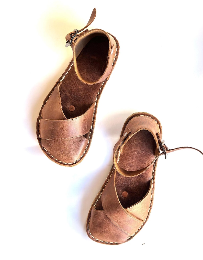 Op de afbeelding: Een paar bruine leren sandalen met een bandje en gespsluiting. De sandalen hebben een kruisvormig ontwerp over de voet en witte stiksels rond de randen. De sandalen worden op een witte achtergrond getoond.