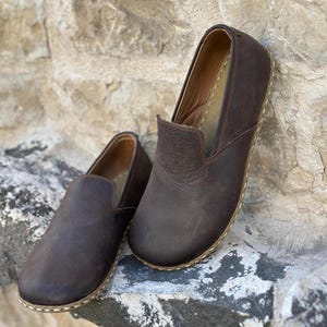 May include: A pair of brown leather slip-on shoes with a stitched edge. The shoes are on a stone surface.