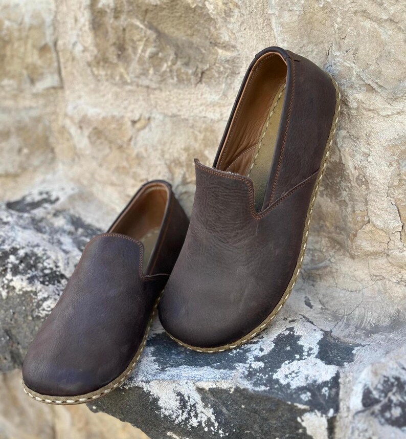 May include: A pair of brown leather slip-on shoes with a stitched edge. The shoes are on a stone surface.