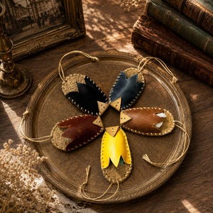 May include: Five oval-shaped leather ornaments arranged in a star pattern on a round tray. Each ornament features a textured base, a smooth petal-shaped overlay in black, dark blue, brown, yellow, or dark red, and a thin hanging cord.
