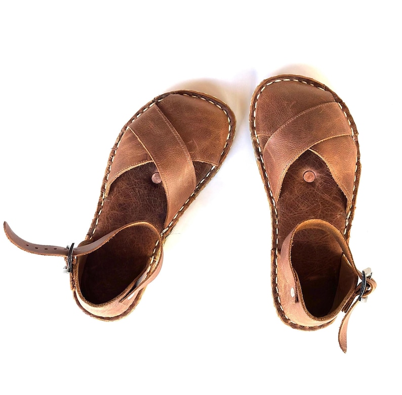 Op de afbeelding: Een paar bruinleren sandalen met een kruisontwerp aan de bovenkant. De sandalen hebben enkelbandjes met gespen en zijn aan de randen met wit garen gestikt. Op een witte achtergrond.