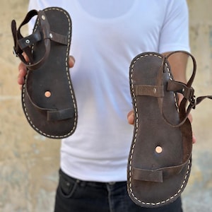 May include: A pair of brown leather sandals with a simple design and a single strap across the foot. The sandals have a white stitching detail around the edges.