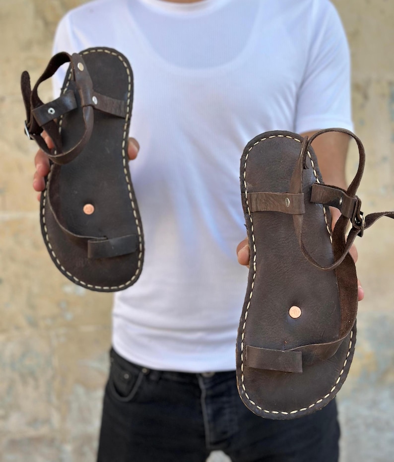 May include: A pair of brown leather sandals with a simple design and a single strap across the foot. The sandals have a white stitching detail around the edges.