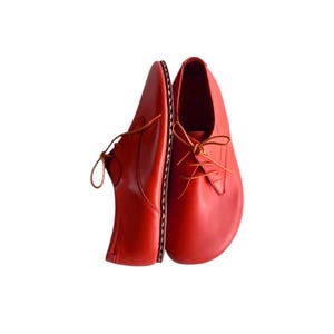 May include: A pair of bright red leather shoes with a rounded toe design and thin matching laces. The shoes feature a visible stitched welt along the side and a smooth, clean finish on the upper surface, presented in a side-by-side orientation.