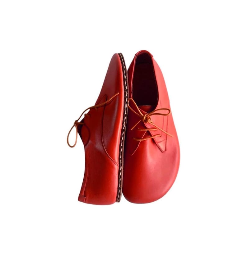 May include: A pair of bright red leather shoes with a rounded toe design and thin matching laces. The shoes feature a visible stitched welt along the side and a smooth, clean finish on the upper surface, presented in a side-by-side orientation.