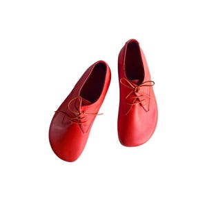 May include: A pair of bright red leather shoes with a rounded toe design and thin matching laces. The shoes feature a low-cut silhouette with a smooth, unpatterned surface and a simple lace-up closure system centered on the top of each shoe.
