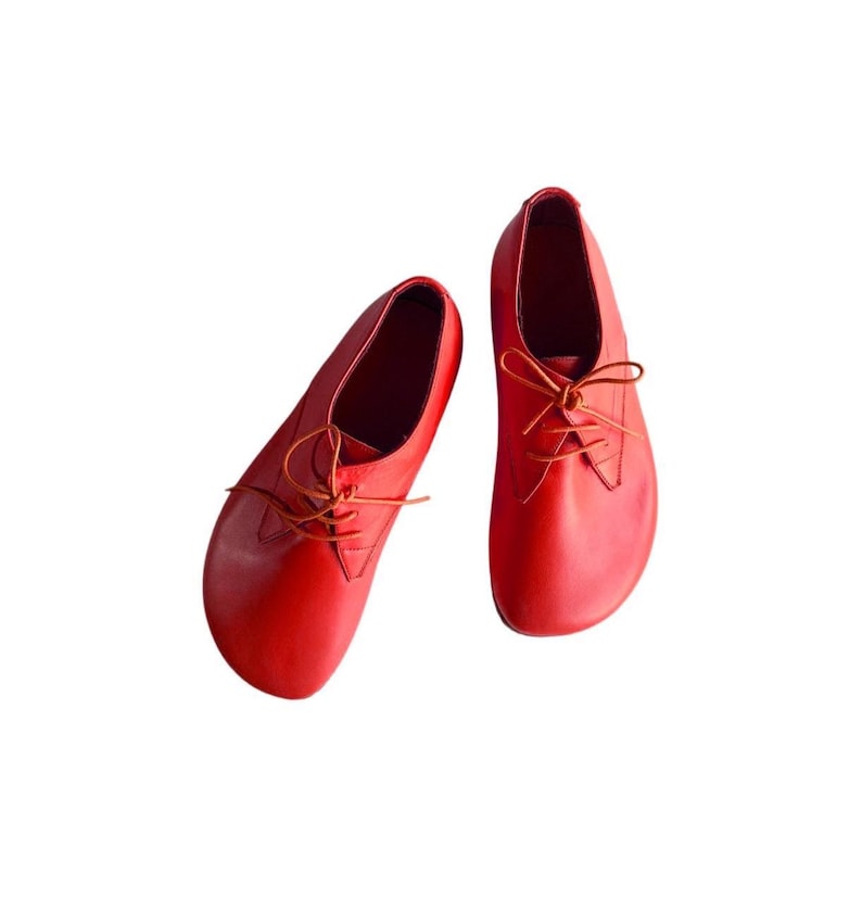 May include: A pair of bright red leather shoes with a rounded toe design and thin matching laces. The shoes feature a low-cut silhouette with a smooth, unpatterned surface and a simple lace-up closure system centered on the top of each shoe.