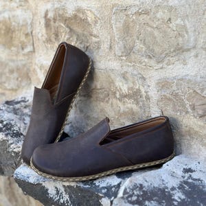 May include: A pair of brown leather slip-on shoes with a white stitching detail around the sole.