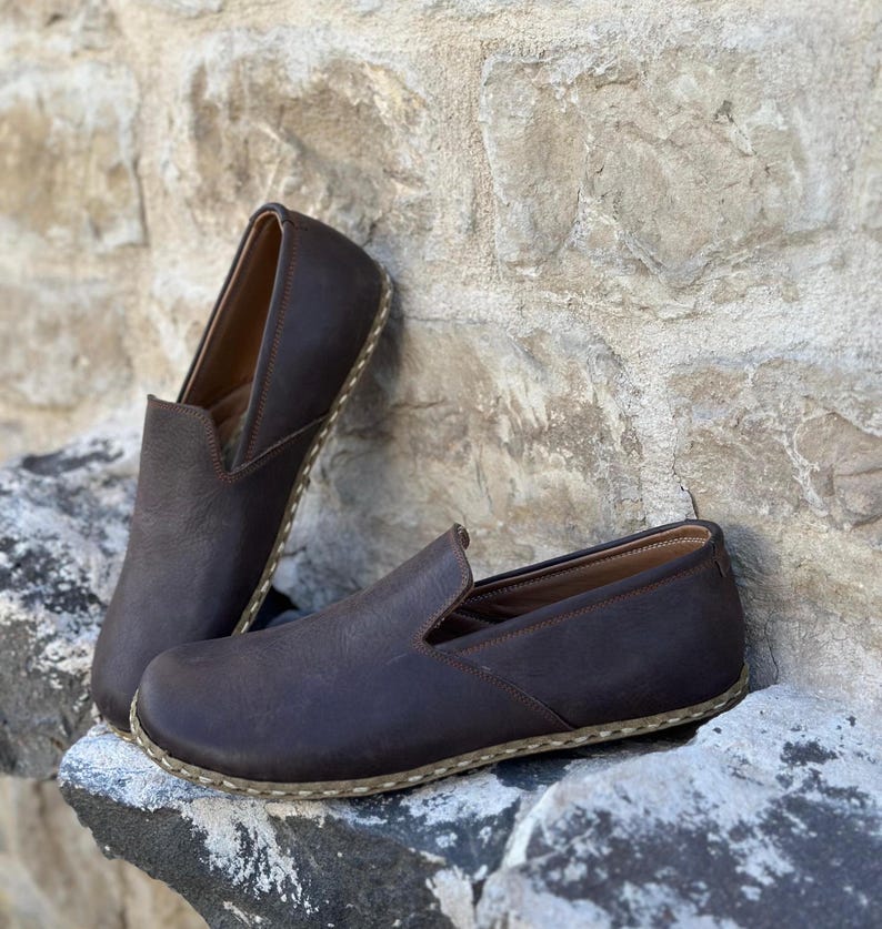 May include: A pair of brown leather slip-on shoes with a white stitching detail around the sole.