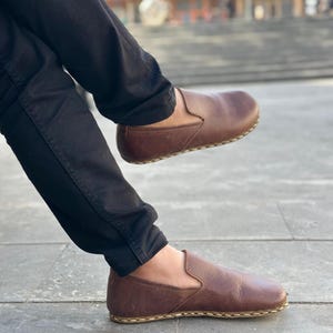 K&ouml;nnte beinhalten: Ein Paar braune Slipper aus Leder mit einem gen&auml;hten Rand. Die Schuhe werden von einer Person mit schwarzer Hose getragen.
