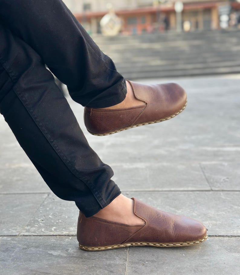 K&ouml;nnte beinhalten: Ein Paar braune Slipper aus Leder mit einem gen&auml;hten Rand. Die Schuhe werden von einer Person mit schwarzer Hose getragen.