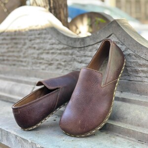 K&ouml;nnte beinhalten: Zwei Paar braune Slipper aus Leder mit einem gen&auml;hten Rand. Die Schuhe befinden sich auf einer Steinoberfl&auml;che.