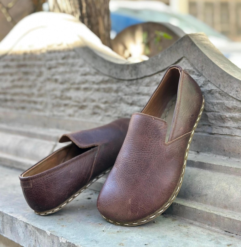 K&ouml;nnte beinhalten: Zwei Paar braune Slipper aus Leder mit einem gen&auml;hten Rand. Die Schuhe befinden sich auf einer Steinoberfl&auml;che.