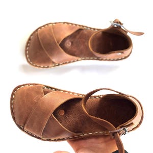 Op de afbeelding: Twee bruine leren sandalen met gekruiste bandjes en enkelbandjes. De sandalen hebben een gestikte rand en een gespsluiting. Het leer heeft een natuurlijk, getextureerd uiterlijk. De sandalen worden op een witte achtergrond getoond.