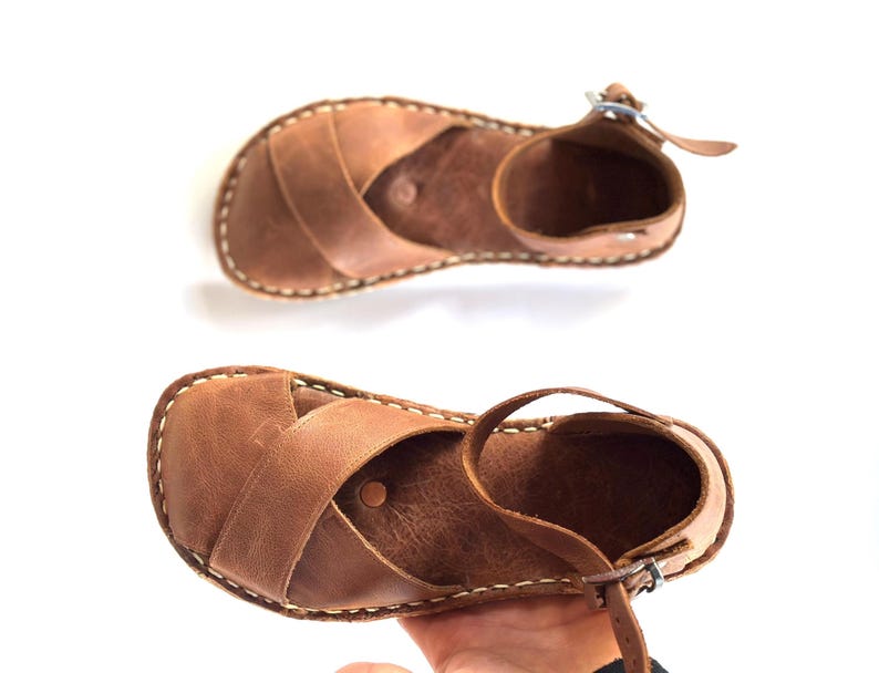 Op de afbeelding: Twee bruine leren sandalen met gekruiste bandjes en enkelbandjes. De sandalen hebben een gestikte rand en een gespsluiting. Het leer heeft een natuurlijk, getextureerd uiterlijk. De sandalen worden op een witte achtergrond getoond.