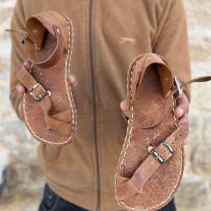 May include: A pair of brown leather sandals with a buckle closure. The sandals have a stitched edge and a textured leather upper.