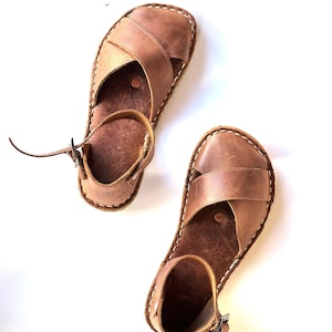 Op de afbeelding: Een paar bruinleren sandalen met een kruisontwerp over de voet en een enkelband met een gesp. De sandalen hebben witte stiksels en staan tegen een witte achtergrond. Zomerschoeisel.