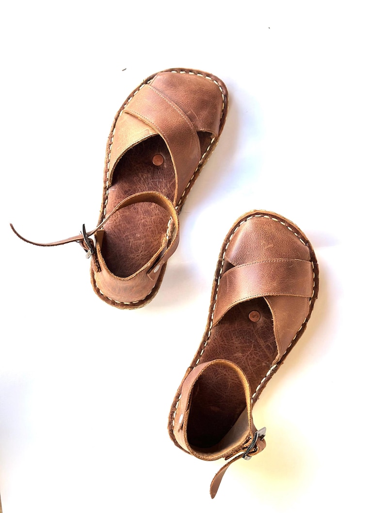 Op de afbeelding: Een paar bruinleren sandalen met een kruisontwerp over de voet en een enkelband met een gesp. De sandalen hebben witte stiksels en staan tegen een witte achtergrond. Zomerschoeisel.