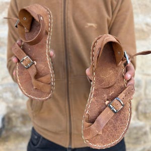 May include: A pair of brown leather sandals with a buckle closure. The sandals have a stitched edge and a distressed leather look.