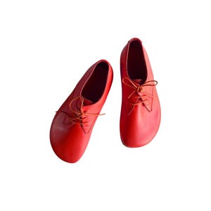 May include: A pair of bright red leather shoes featuring a rounded toe design, a low-cut silhouette, and thin matching red laces tied in a bow. The shoes have a smooth, matte finish and a simple construction with visible stitching along the eyelets.