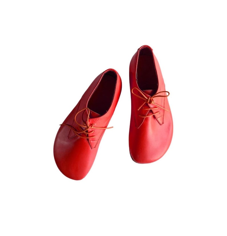 May include: A pair of bright red leather shoes featuring a rounded toe design, a low-cut silhouette, and thin matching red laces tied in a bow. The shoes have a smooth, matte finish and a simple construction with visible stitching along the eyelets.