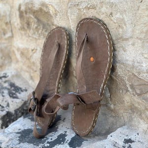 Puede incluir: Un par de sandalias de cuero marr&oacute;n con una correa de estilo thong y un borde cosido. Las sandalias tienen un cierre de hebilla en la correa del tobillo.