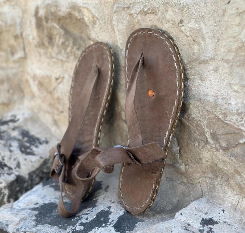 Puede incluir: Un par de sandalias de cuero marr&oacute;n con una correa de estilo thong y un borde cosido. Las sandalias tienen un cierre de hebilla en la correa del tobillo.
