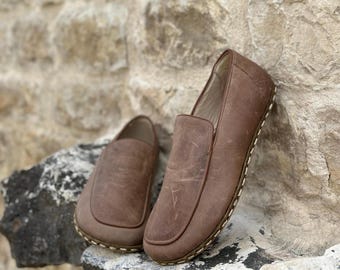 May include: Two brown leather slip-on shoes with white stitching around the edges. The shoes are on a rock surface.