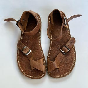 Puede incluir: Un par de sandalias de cuero marrón con correas en el tobillo y presillas para los dedos. Las sandalias tienen hebillas plateadas y costuras blancas alrededor de los bordes. El cuero tiene una textura.