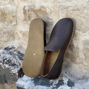 May include: A pair of brown leather slip-on shoes with a stitched edge. The shoes are on a stone surface.