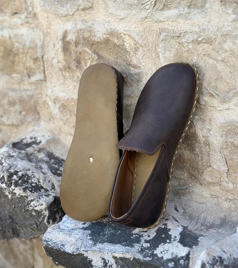 May include: A pair of brown leather slip-on shoes with a stitched edge. The shoes are on a stone surface.