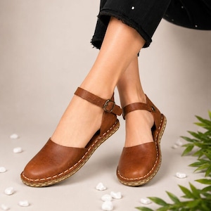 Peut inclure: Une paire de chaussures plates en cuir marron avec un bout arrondi, une bride de cheville dotée d'une boucle métallique et des coutures apparentes de couleur claire le long de la semelle. Le modèle présente des côtés ouverts et un talon plat.