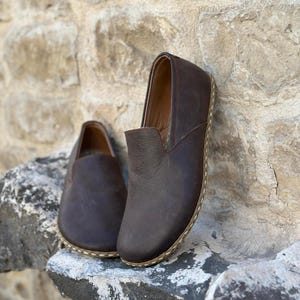 May include: A pair of brown leather slip-on shoes with a stitched edge detail. The shoes are on a stone surface.