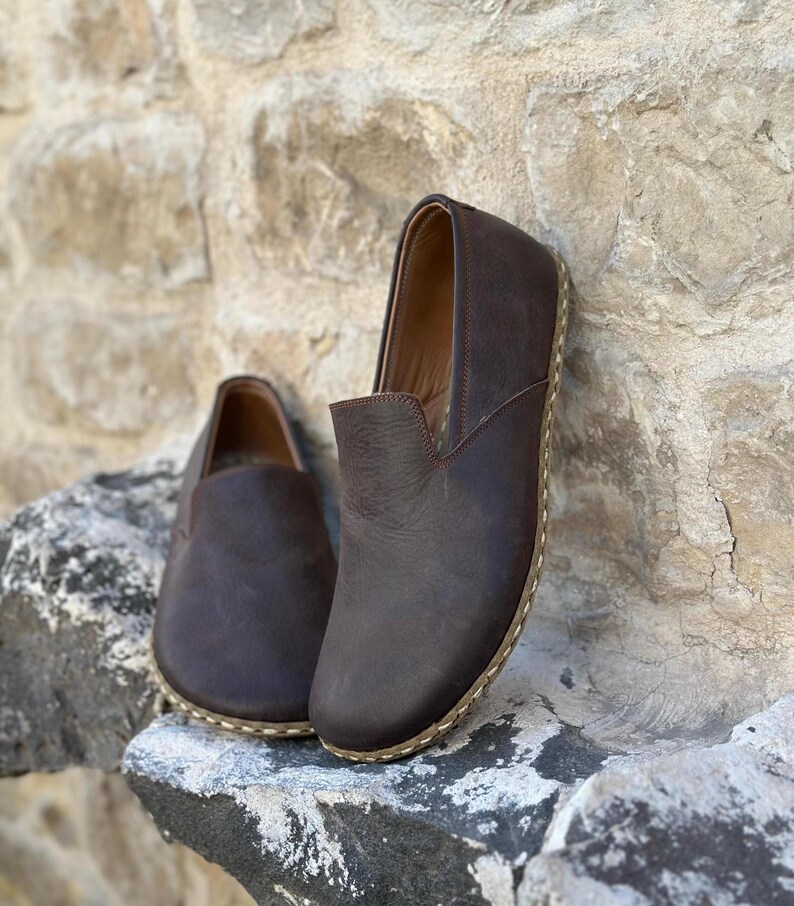 May include: A pair of brown leather slip-on shoes with a stitched edge detail. The shoes are on a stone surface.