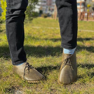 May include: A pair of brown leather lace-up shoes with a rounded toe. The shoes are worn by a person wearing black jeans and light blue socks.