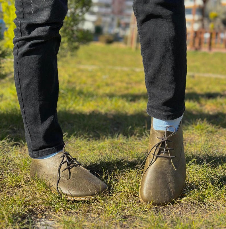 May include: A pair of brown leather lace-up shoes with a rounded toe. The shoes are worn by a person wearing black jeans and light blue socks.