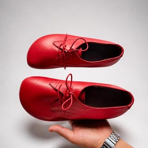May include: A pair of bright red leather shoes with a rounded toe box and a lace-up closure. The shoes feature a smooth finish, thin matching red laces, and a low-cut ankle opening. The design includes a wide forefoot area and a simple construction.