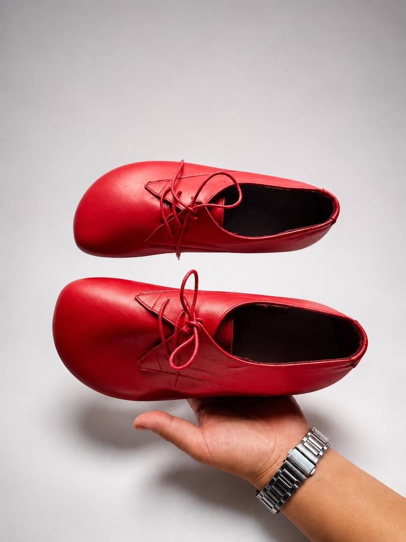 May include: A pair of bright red leather shoes with a rounded toe box and a lace-up closure. The shoes feature a smooth finish, thin matching red laces, and a low-cut ankle opening. The design includes a wide forefoot area and a simple construction.
