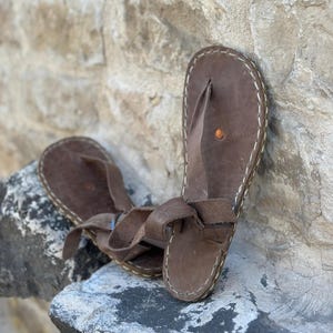 Puede incluir: Un par de sandalias de cuero marr&oacute;n con un dise&ntilde;o sencillo. Las sandalias tienen una sola correa que va alrededor del tobillo y un bucle para los dedos. Las sandalias est&aacute;n hechas de cuero marr&oacute;n y tienen costuras blancas alrededor de los bordes.