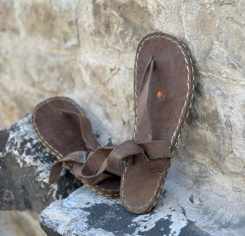 Puede incluir: Un par de sandalias de cuero marr&oacute;n con un dise&ntilde;o sencillo. Las sandalias tienen una sola correa que va alrededor del tobillo y un bucle para los dedos. Las sandalias est&aacute;n hechas de cuero marr&oacute;n y tienen costuras blancas alrededor de los bordes.