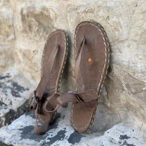 Puede incluir: Un par de sandalias de cuero marr&oacute;n con una correa de estilo thong y un detalle de costura blanca alrededor de los bordes. Las sandalias tienen un cierre de hebilla en la correa del tobillo.