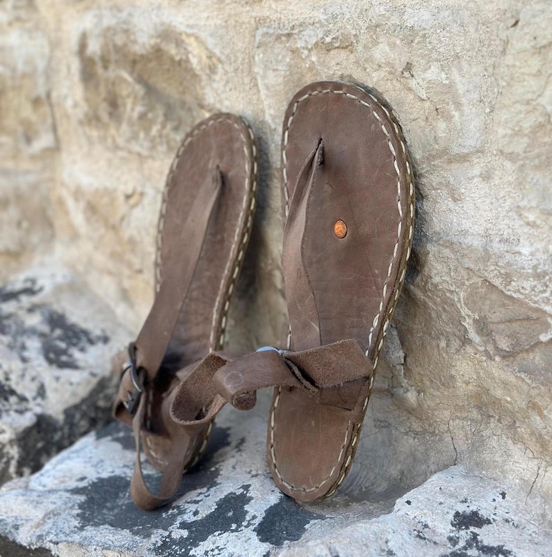 Puede incluir: Un par de sandalias de cuero marr&oacute;n con una correa de estilo thong y un detalle de costura blanca alrededor de los bordes. Las sandalias tienen un cierre de hebilla en la correa del tobillo.