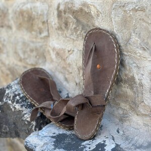 Puede incluir: Un par de sandalias de cuero marr&oacute;n con un detalle de costura blanca alrededor de los bordes. Las sandalias tienen un dise&ntilde;o sencillo con una sola correa sobre el pie y un bucle para el dedo del pie.