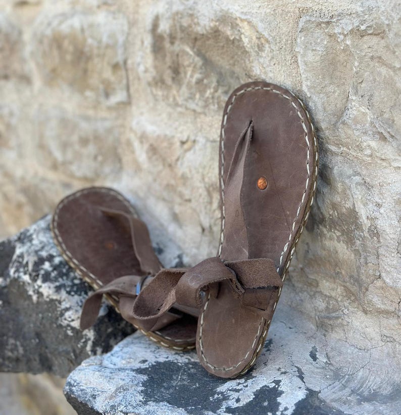 Puede incluir: Un par de sandalias de cuero marr&oacute;n con un detalle de costura blanca alrededor de los bordes. Las sandalias tienen un dise&ntilde;o sencillo con una sola correa sobre el pie y un bucle para el dedo del pie.