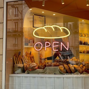 May include: A bakery storefront with a neon sign that says "OPEN" in red. Above the text is a yellow neon outline of a loaf of bread. Various loaves of bread are displayed behind the glass window.