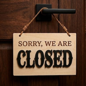 Op de afbeelding: Een rechthoekig, lichtgekleurd houten bordje dat aan een zwarte deurklink hangt met een beige leren koord. Het bordje bevat de tekst SORRY, WE ARE in een eenvoudig lettertype en het woord CLOSED in grote, dikke, zwarte letters.